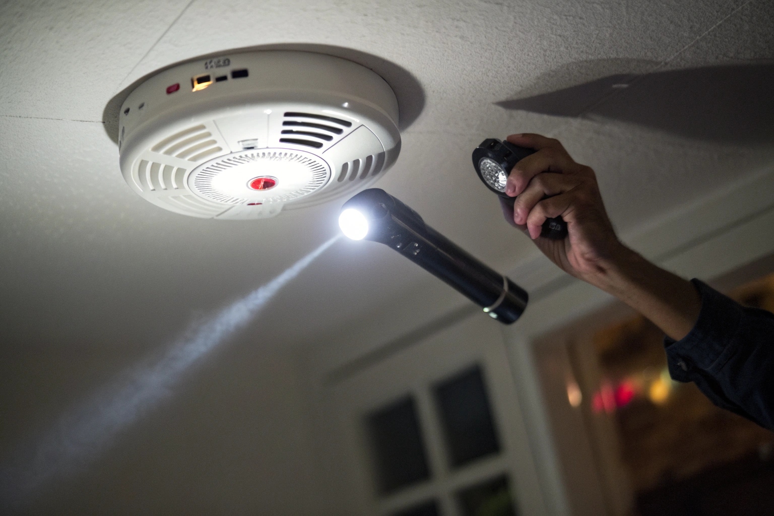 Gros plan d'une main utilisant une lampe torche pour révéler le reflet d'un minuscule objectif d'appareil photo dissimulé dans un détecteur de fumée